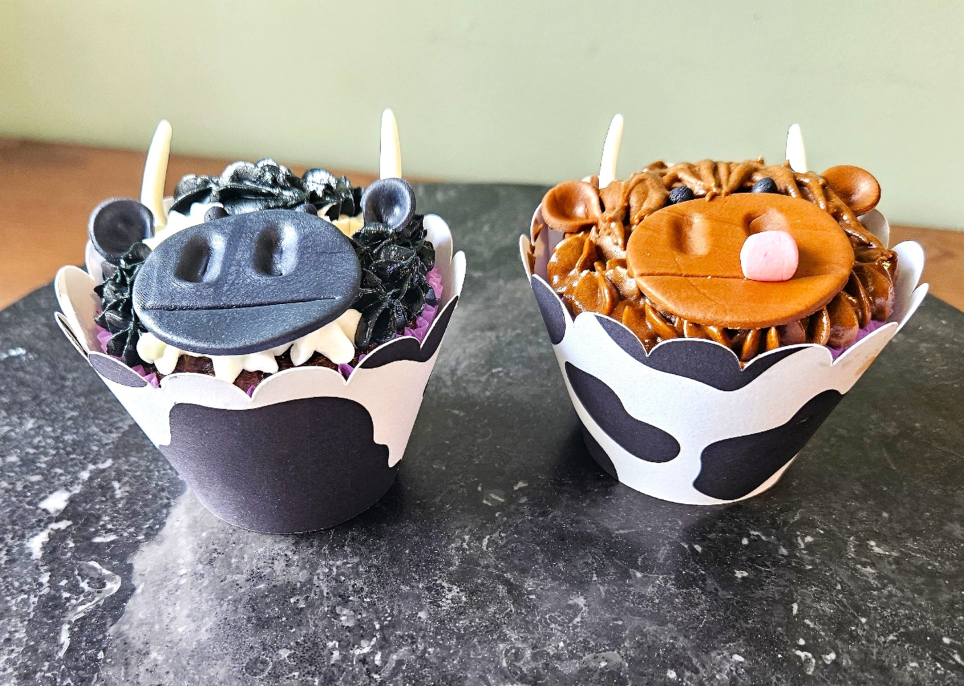 Two cupcakes with cow-themed decorations on a dark surface.