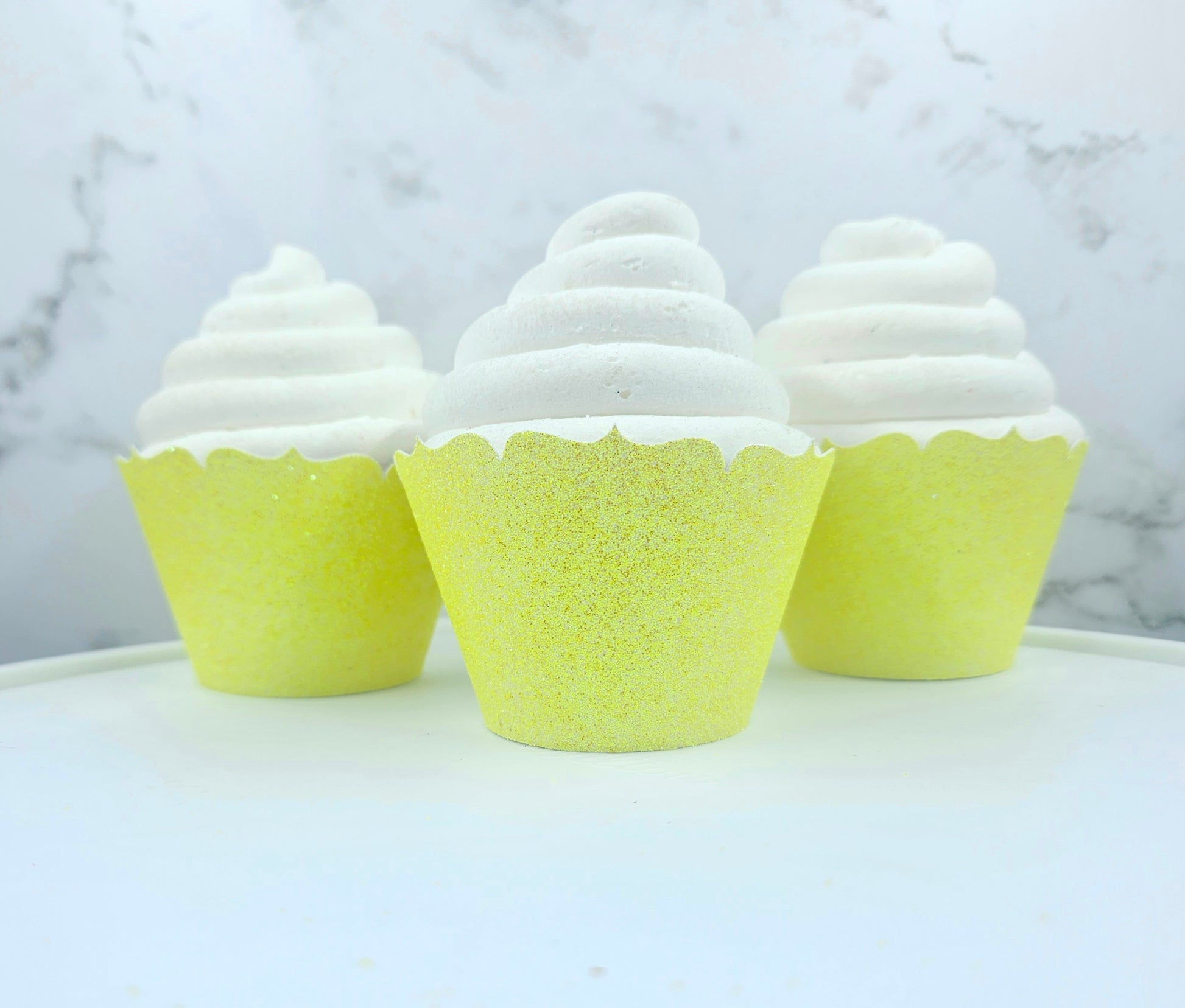 Three cupcakes with white frosting and yellow cupcake liners on a marble surface.